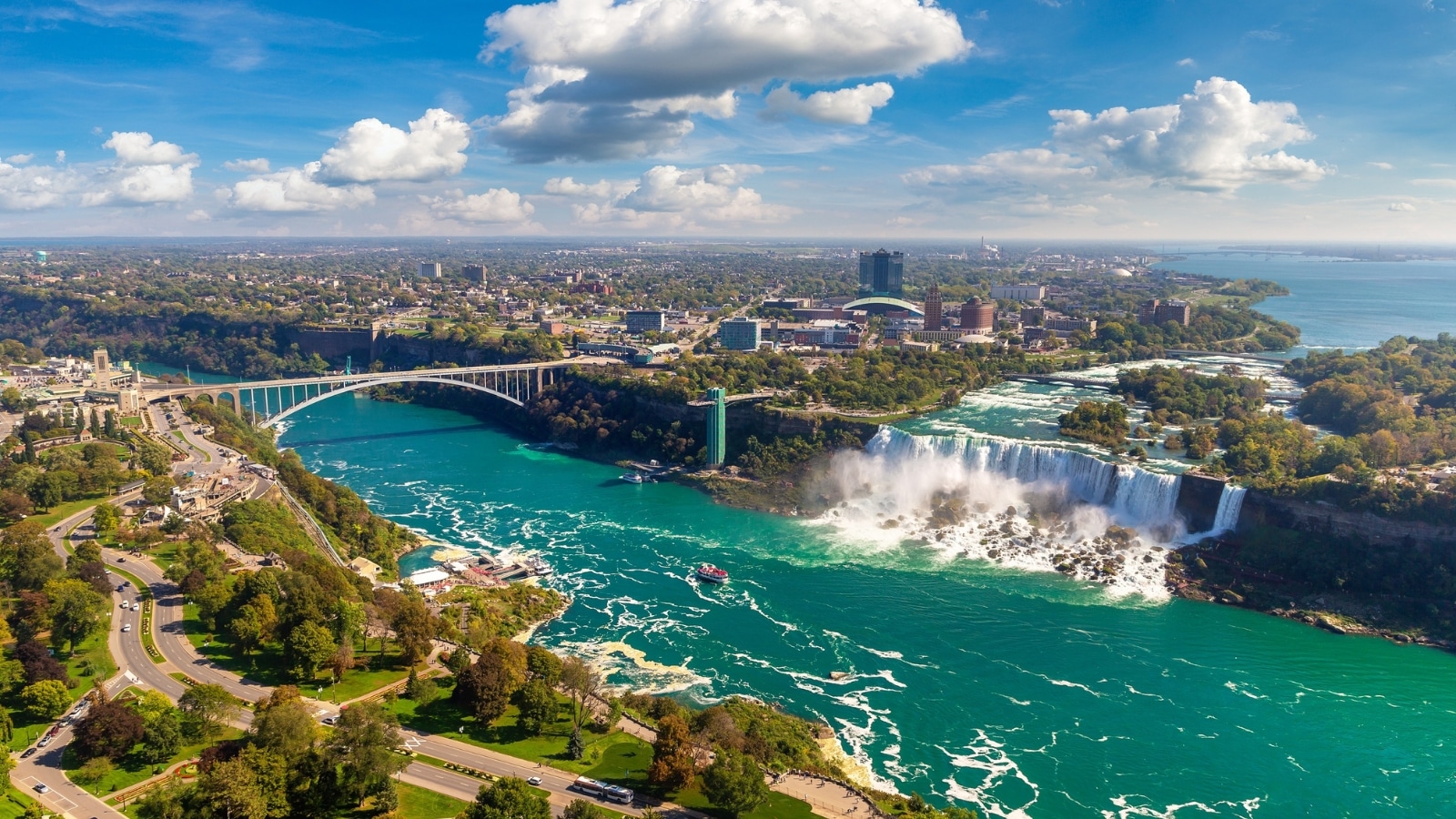 The image displays a panoramic aerial view of the Canadian side of Niagara Falls, including the American Falls and the Rainbow International Bridge, all under a sunny sky in Niagara Falls, Ontario, Canada.