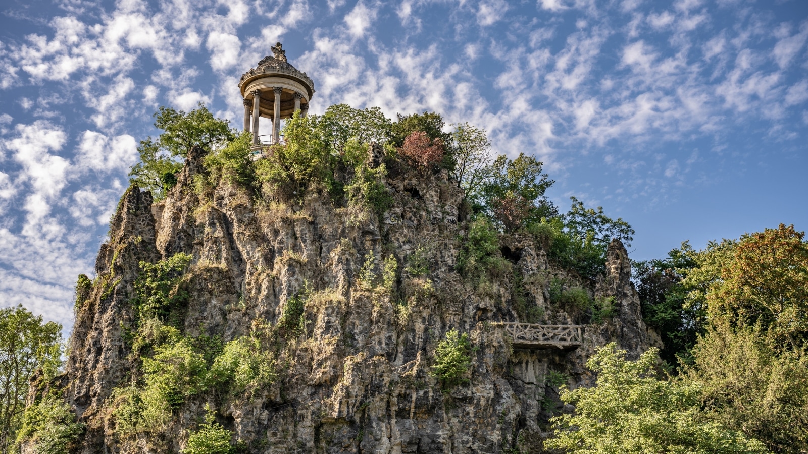 The image shows a view of Parc des Buttes-Chaumont in Paris, France. A prominent feature is a large rock adorned with greenery, atop which sits an observation deck supported by columns and a roof.