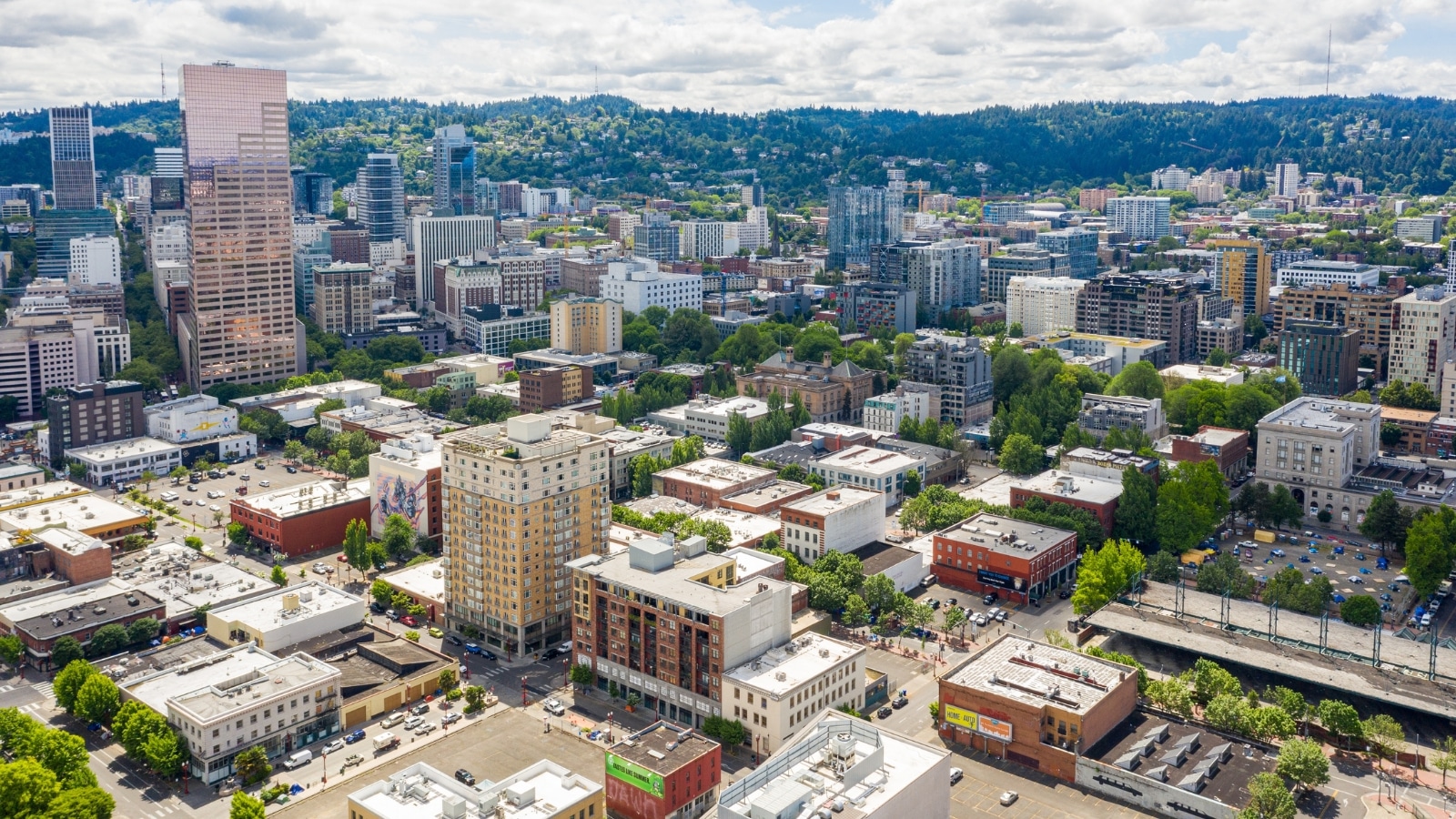 The image provides an aerial perspective of Downtown Portland, Oregon, showcasing a mix of 1-3 story buildings along with high-rise structures. The streets and roads are clearly defined, illustrating the urban layout of the area. Lining the streets are numerous green trees, contributing to the city's greenery and aesthetics. In the background, small hills can be seen.