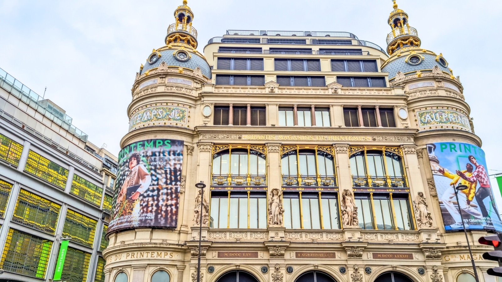 The image captures a view of the Printemps department store in Paris, France. The building features a blend of modern and classic architectural elements, with ornate details and a grand facade. The color scheme includes shades of cream, beige, and gold, typical of historic Parisian architecture.