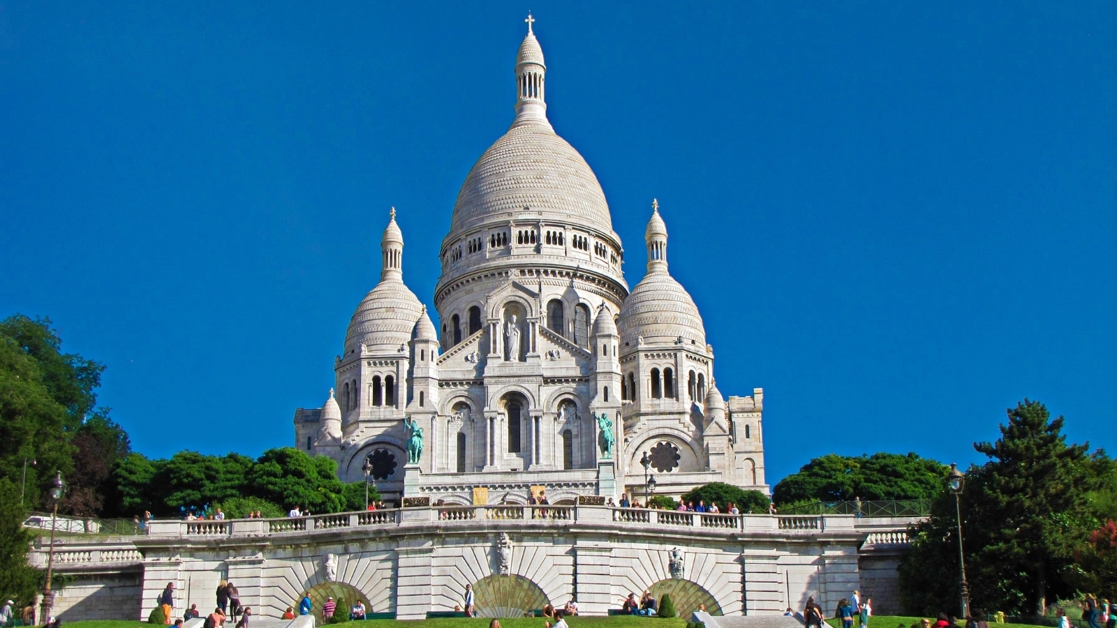 The image captures a scene in Paris, France, focusing on the Sacre Coeur Basilica. Surrounding the basilica, there is a significant amount of greenery, including a trimmed lawn, adding to the aesthetic appeal of the area. Numerous people can be seen walking and climbing the steps leading to the basilica. The Sacre Coeur Basilica itself is notable for its white exterior and features three domes, with the central dome being the largest and most prominent.