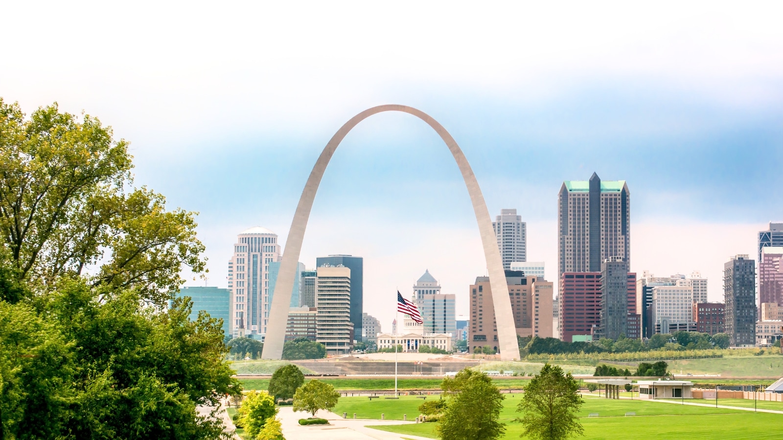 The image depicts the St. Louis skyline with a prominent display of the American flag positioned under the iconic Gateway Arch. The sky appears overcast. Beneath the arch, there is a well-maintained flat lawn and several trees. Beyond the arch, the cityscape unfolds with tall buildings.