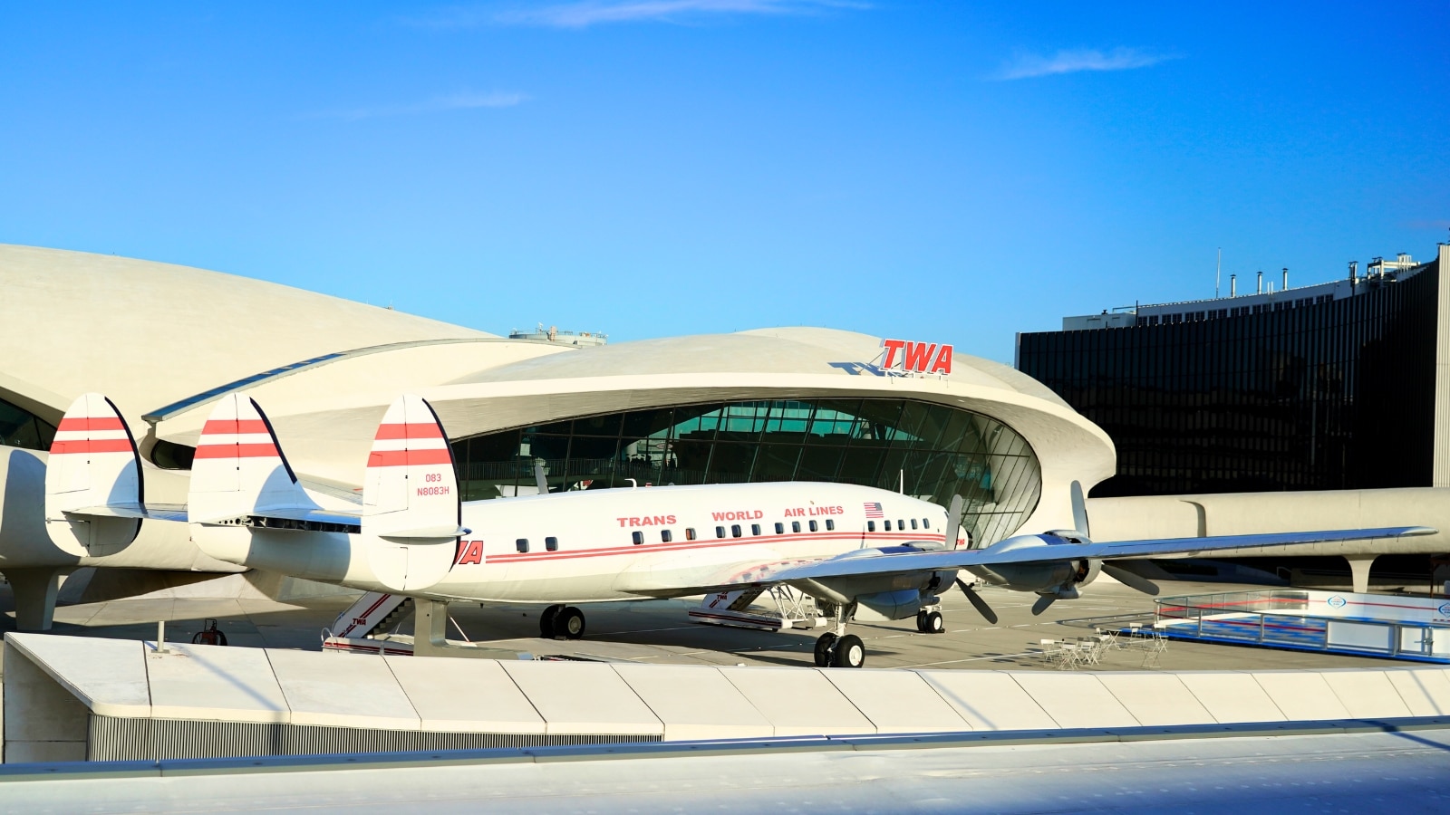 The image features the TWA Hotel at New York's John F. Kennedy Airport, characterized by its futuristic white architecture prominently displaying the TWA sign in red letters. In the foreground, a white plane with red stripes is parked in front of the building, adding to the aviation-themed ambiance of the hotel.