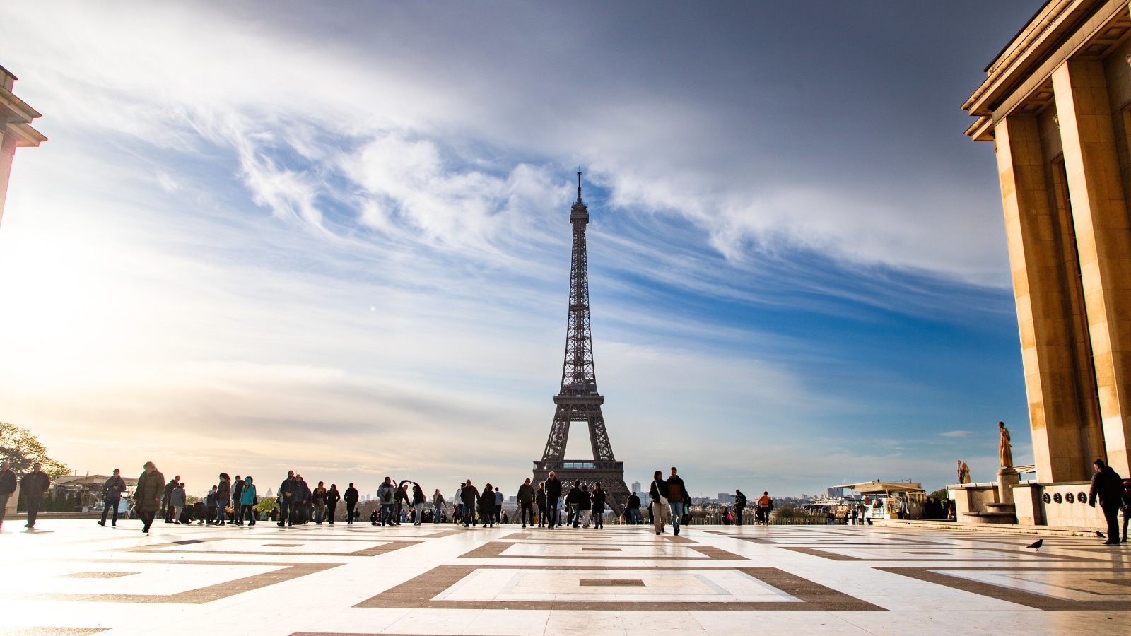 The location described is the view of the Eiffel Tower from the Jardins du Trocadéro in Paris, France. The perspective is from ground level, where the floor is tiled with square patterns. Many people can be seen observing the Eiffel Tower from this vantage point.