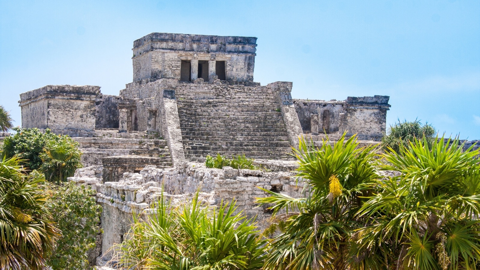 The image depicts the Tulum Ruins, an ancient Mayan archaeological site located along the eastern coast of the Yucatán Peninsula in Mexico. The ruins are characterized by well-preserved stone structures, including temples.