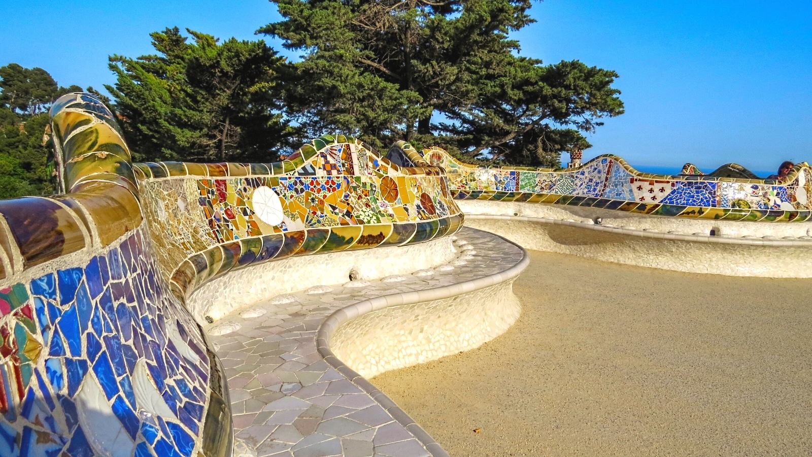 The image captures a view of a mosaic tiled sitting bench located in a park in Barcelona, Spain. The bench's design features intricate mosaic patterns, showcasing artistic craftsmanship.