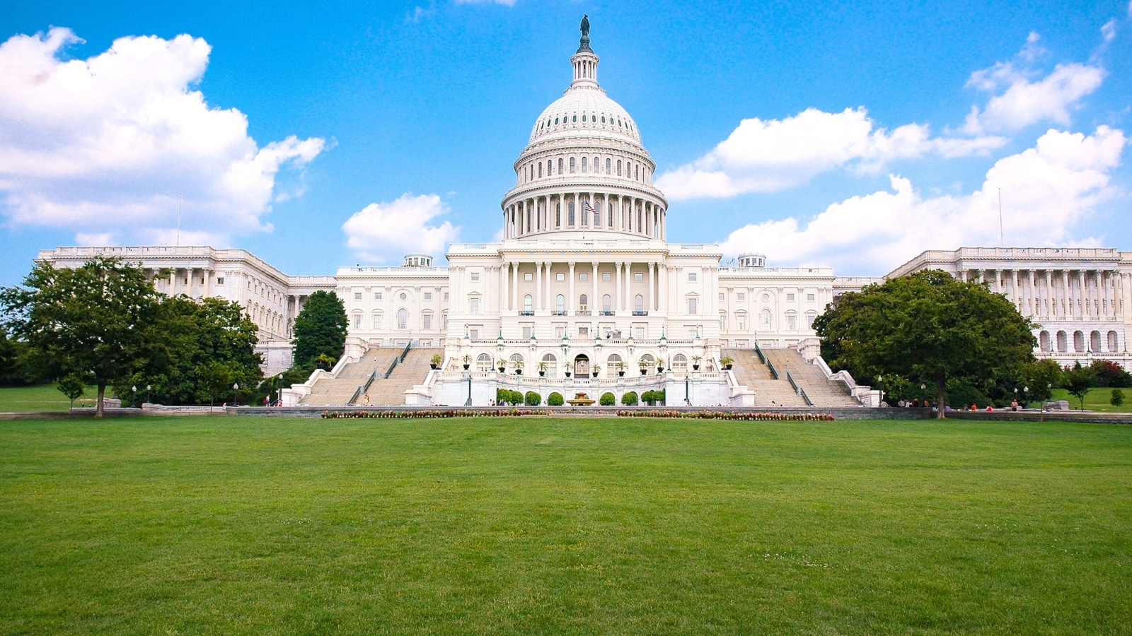 The image features the United States Capitol Building in Washington, D.C., against a backdrop of clear skies on a sunny day. In front of the building, there is a well-maintained green lawn and several trees. The white façade of the building stands out against the blue sky, creating a visually contrast.