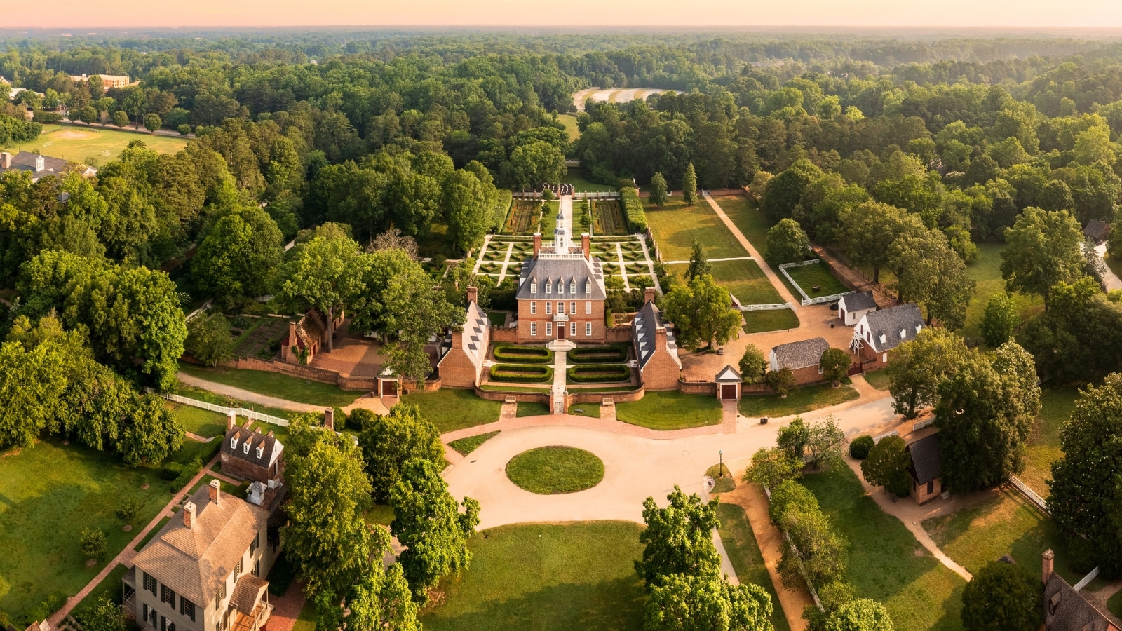 The image offers a drone view of the front facade of the Governors Palace in Colonial Williamsburg, Virginia, captured at dawn. Surrounding the palace are extensive greenery and trees.