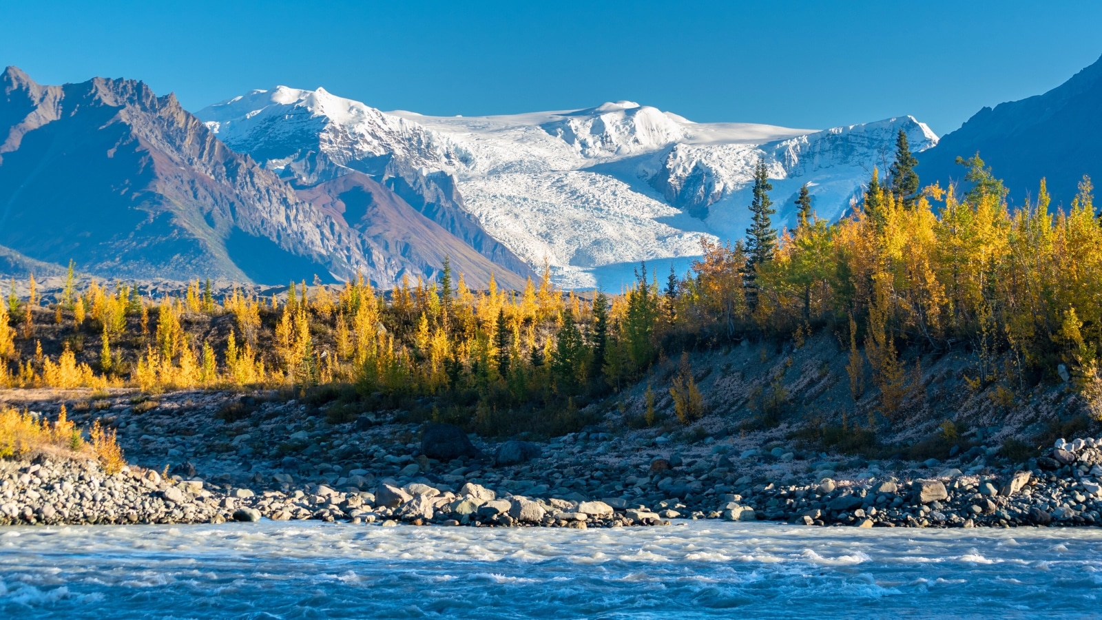 Wrangell-St. Elias National Park & Preserve, featuring a river in the foreground, a hilly stone bank slightly elevated, and snow-capped mountains in the background.