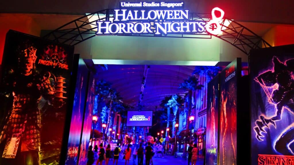 The image captures the entrance to Universal Studio's Halloween Horror Nights, set in a dark environment illuminated by neon signs. The scene is eerie, with shadows dominating the surroundings. Neon lights spell out spooky messages, adding to the ominous ambiance.