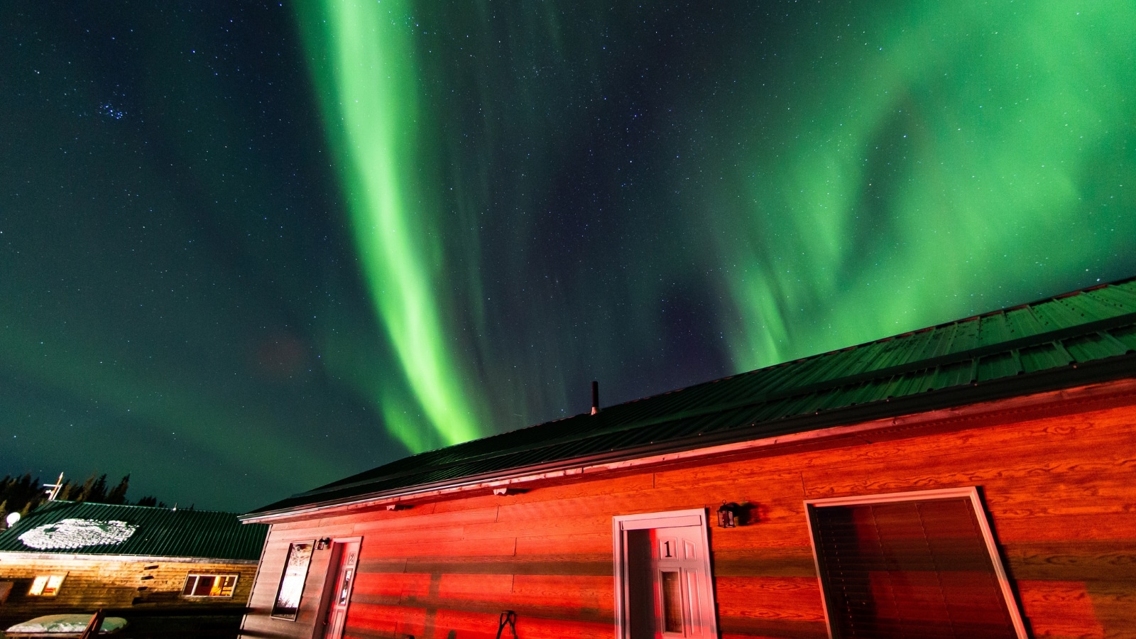 The image showcases a beautiful aurora in Fairbanks, Alaska, with a one-story building in the foreground illuminated in red tones. In the background, the sky displays a green glow, typical of auroras.