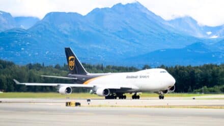 The image depicts a UPS Airlines plane taking off from Anchorage International Airport. The plane is predominantly white with a black livery. In the background, prominent mountains with snow-capped peaks can be seen.