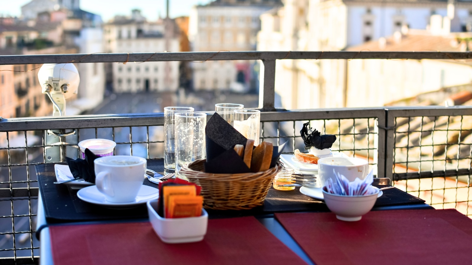 The image depicts the remains of breakfast on a cafe balcony overlooking a blurred square.