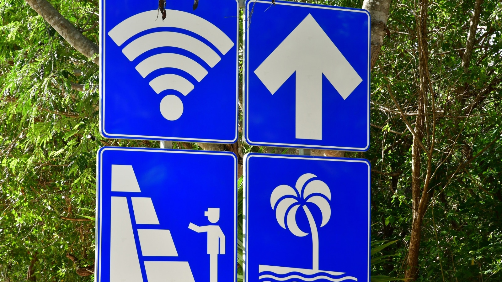 The image is a close-up of a sign in Tulum, Mexico, featuring four blue squares with distinct symbols. One square displays a Wi-Fi icon, indicating the availability of wireless internet. Another square shows an arrow, likely providing directional information. The third square depicts a person next to a pyramid, possibly indicating a tourist attraction or historical site. The fourth square features a palm tree, suggesting tropical surroundings.