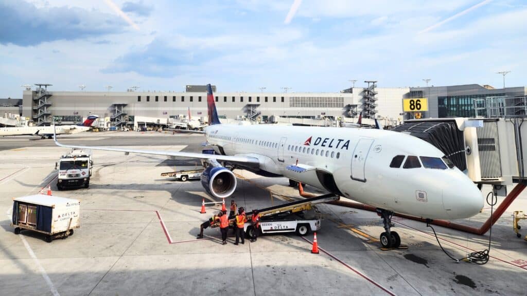 The image depicts a Delta Airlines passenger plane at LaGuardia Airport terminal, preparing for departure. The plane is positioned on the tarmac, surrounded by airport infrastructure.