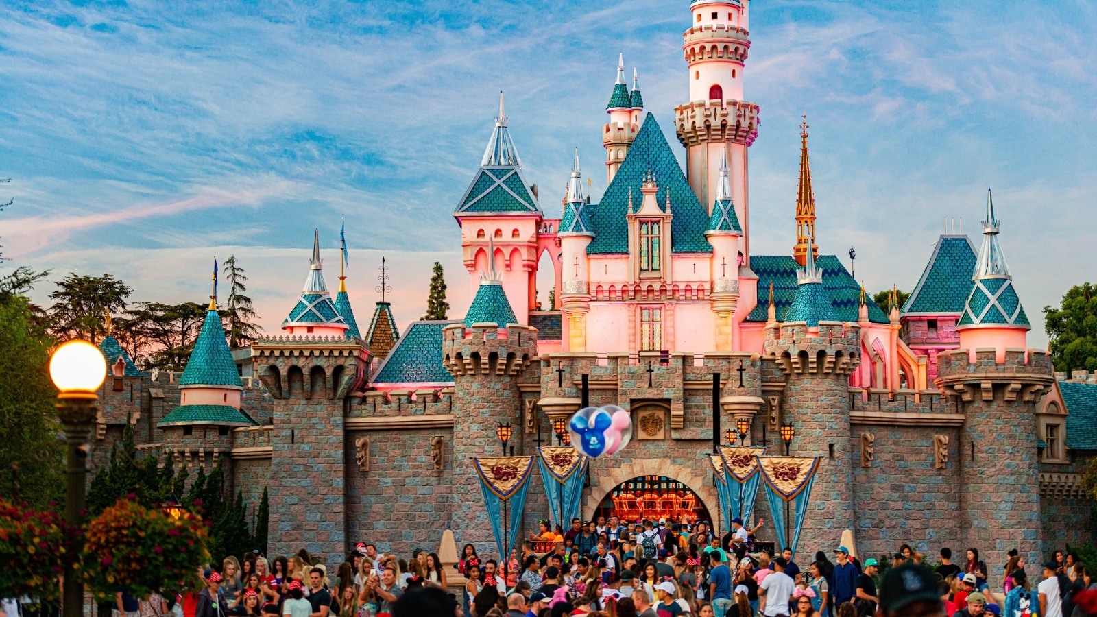 View of the legendary Disney castle of Sleeping Beauty in Disneyland. Its facade is adorned with intricate details and whimsical touches, featuring pastel hues and shimmering accents that evoke fairy tale fantasies. Turrets and towers rise gracefully, crowned with regal flags fluttering in the breeze, while lush gardens and sparkling moats add to the enchanting ambiance. Crowds of people stand at the entrance to the castle.
