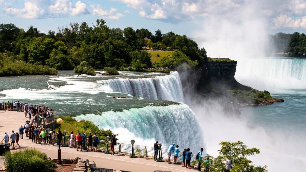 The image captures Niagara Falls State Park, located in New York. This park is characterized by its breathtaking natural beauty and powerful cascades. Towering waterfalls, including the iconic Horseshoe Falls, American Falls, and Bridal Veil Falls, dominate the landscape, sending mist billowing into the air. Lush greenery surrounds the falls, with manicured gardens, scenic overlooks, and winding pathways. Many people stand at the fence watching the waterfalls.