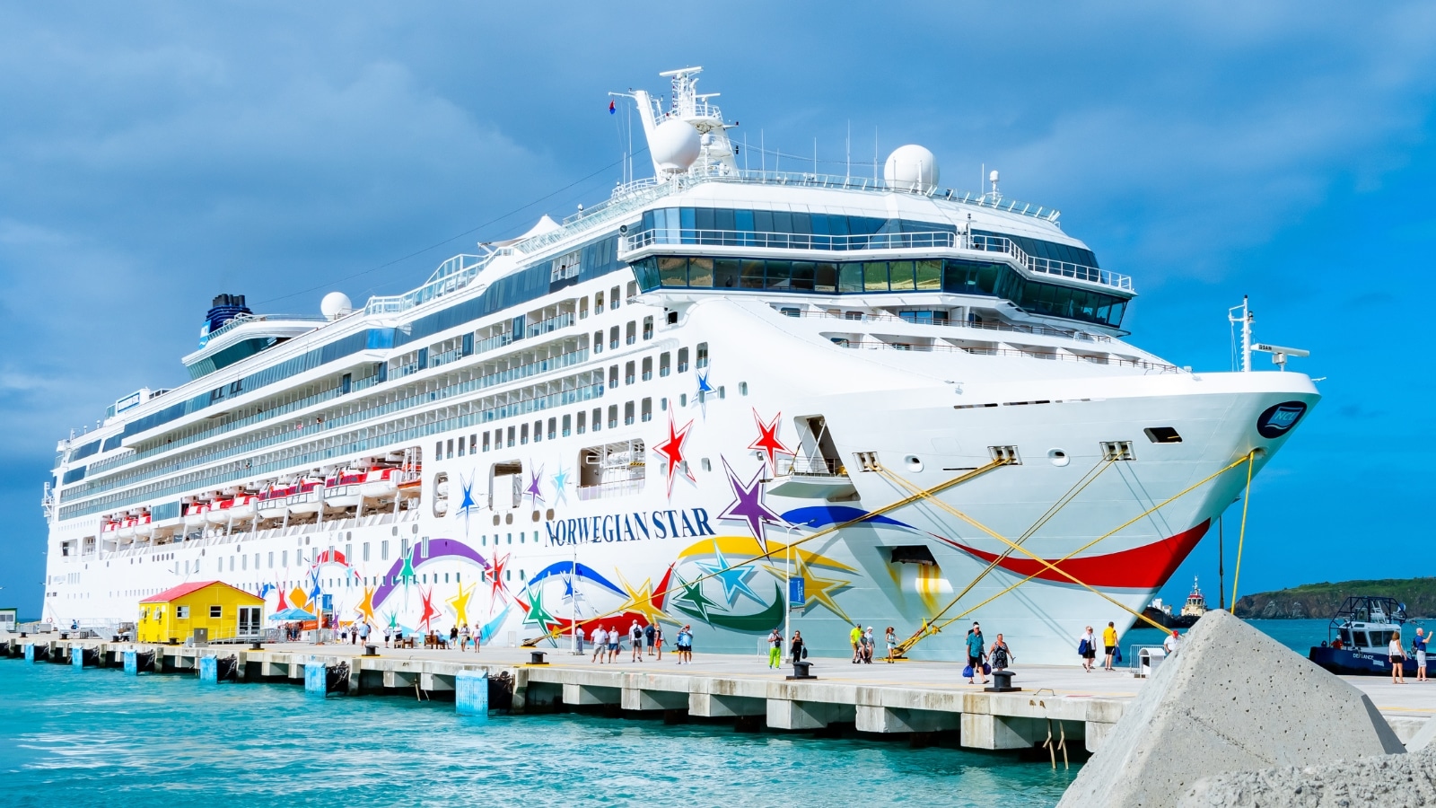 In the image, the Norwegian Star cruise ship is docked at the Sint Maarten port. The ship appears white with vibrant drawings adorning the lower part, prominently displaying the name "Norwegian Star."