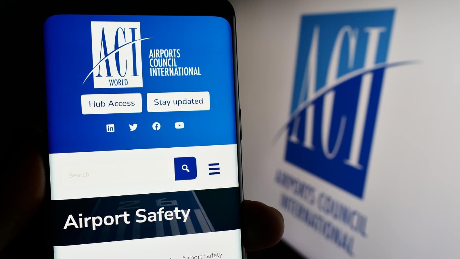The image depicts a person holding a cellphone with the webpage of the organization Airports Council International (ACI) displayed in front of the ACI logo.