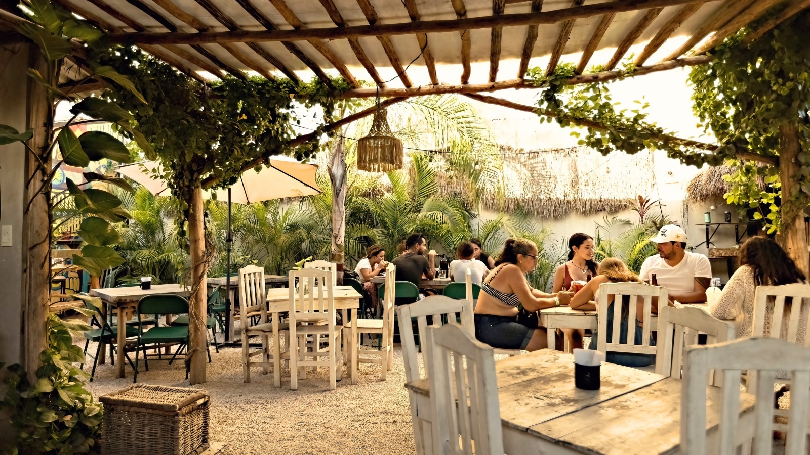 The image depicts people seated at an open-air cafe in Tulum, Mexico, with white tables and chairs. The setting exudes a relaxed and tropical atmosphere.