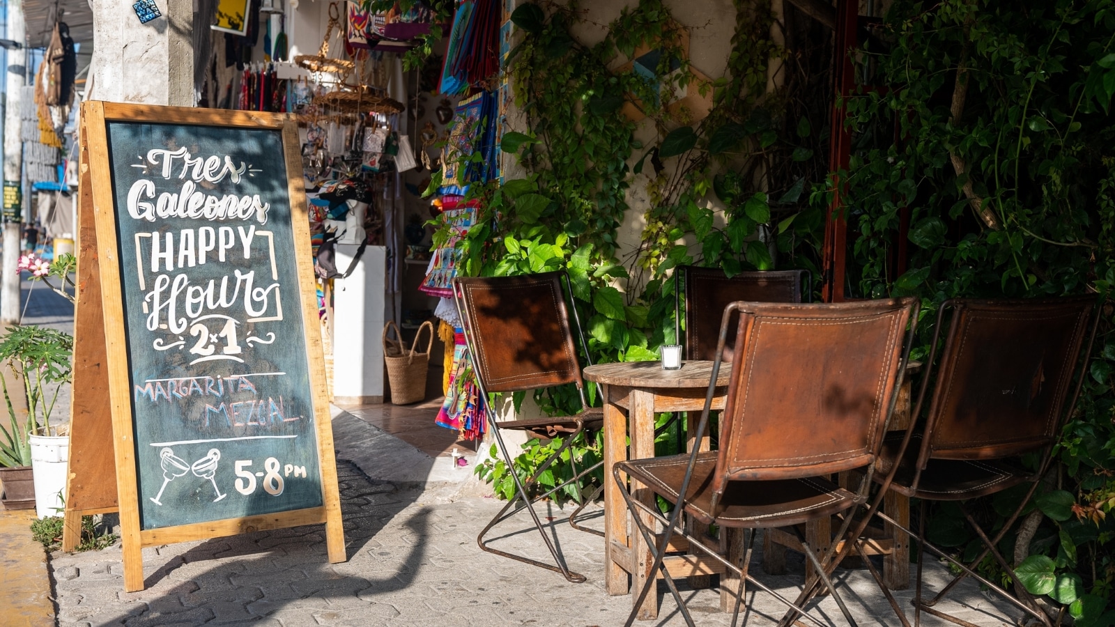 The image features a small establishment in Tulum, Mexico, with chairs and tables arranged outside on the street. Adjacent to the establishment is a board displaying the inscription "Happy Hours."