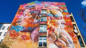 The image displays a decorative mural painting on a skyscraper located in Berlin-Kreuzberg, Germany. The mural consists of graffiti in bright, colorful colors, and prominently features two faces.