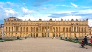 The image features people visiting the Palace of Versailles, a renowned royal chateau located in Versailles, France. Visitors can be seen exploring the expansive grounds. The palace