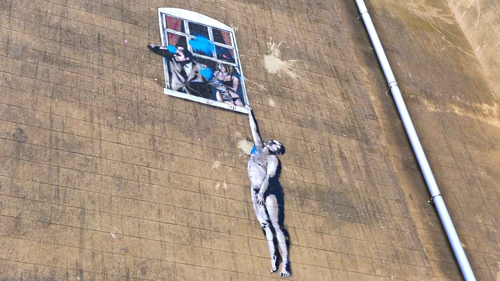 The image captures a graffiti artwork in Bristol, United Kingdom, created by the artist Banksy. Known as "Wall Hanger," the artwork depicts a naked man hanging onto a window sill.