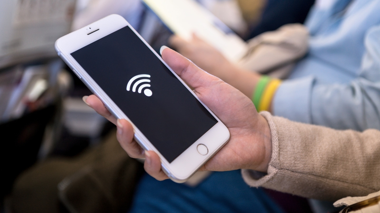 close-up of a woman's hand holding a smartphone with a Wi-Fi logo displayed on the screen. The setting indicates that the woman is seated on a plane, likely accessing in-flight Wi-Fi services.