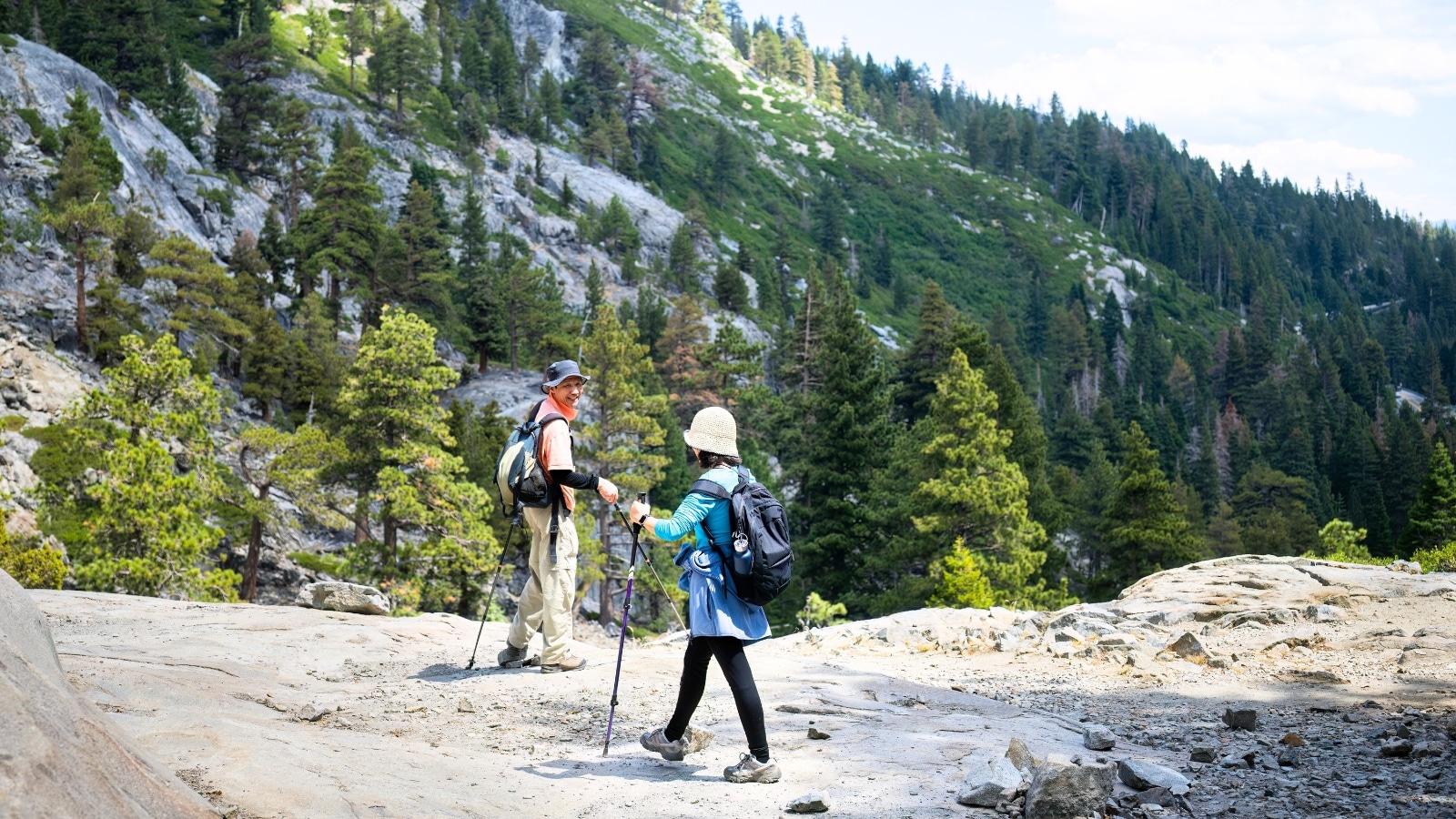 Сouple hiking the Eagle Lake trail in Emerald Bay, South Lake Tahoe, California. Both individuals are wearing bandanas on their heads, carrying backpacks, and holding trekking poles. In the background, lush green mountains surround the trail, typical of the scenic beauty found in the area.