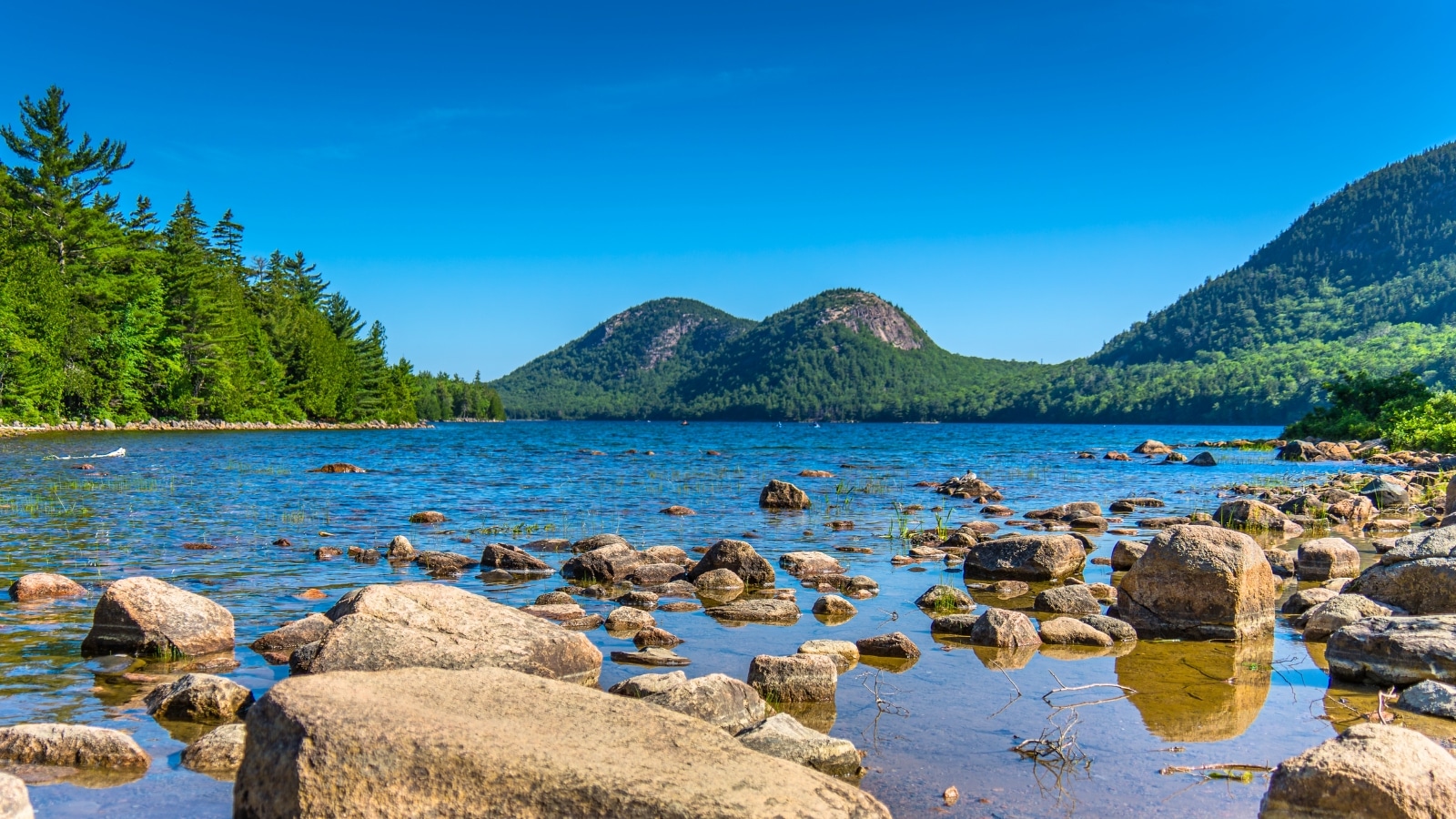 Acadia National Park in Maine features a serene scene with a river in the foreground and rocky terrain nearby. Beyond, green hills rise in the background, adding depth to the landscape.