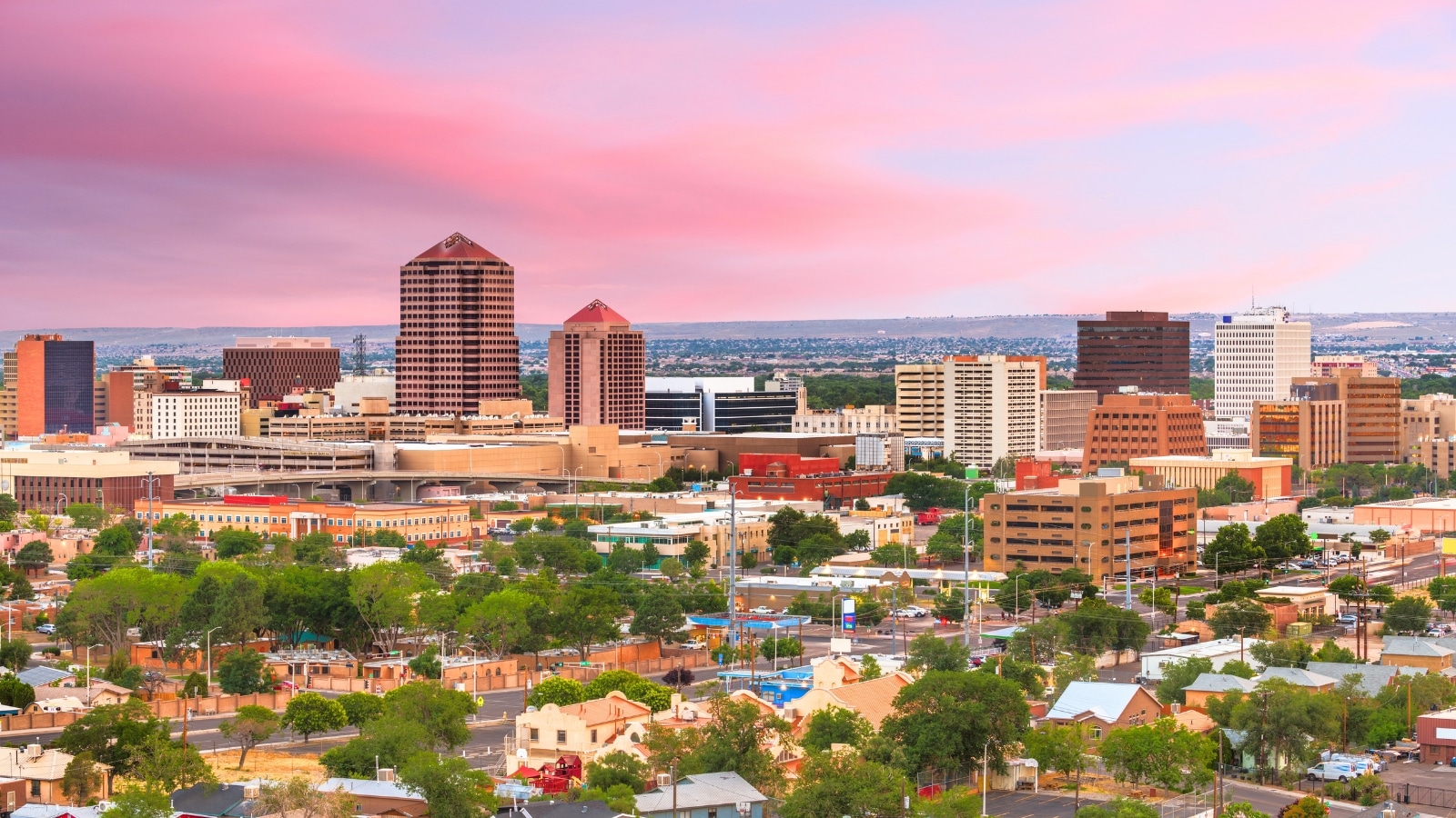 Albuquerque, New Mexico, USA, downtown cityscape at twilight features low buildings in the foreground surrounded by greenery and trees. In the background, the city rises with high buildings, illuminated against the twilight sky.