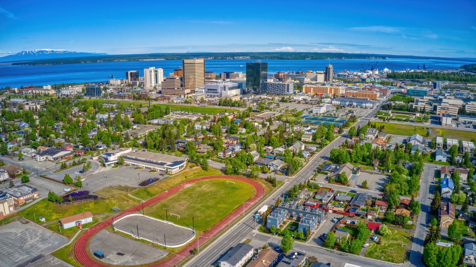 An aerial view captures the downtown Anchorage, Alaska skyline during summer. In the background, a wide river flows with mountains showcasing snow-capped peaks. The foreground features the cityscape with buildings and urban infrastructure.