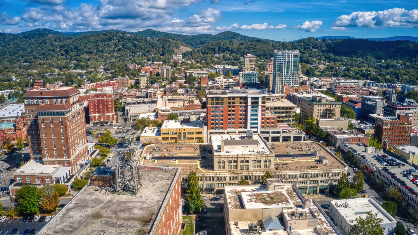 An aerial view of Asheville, North Carolina during summer reveals a vibrant cityscape surrounded by lush greenery. The background is dominated by hills and mountains, densely dotted with trees. The urban area features a mix of modern and historic buildings.