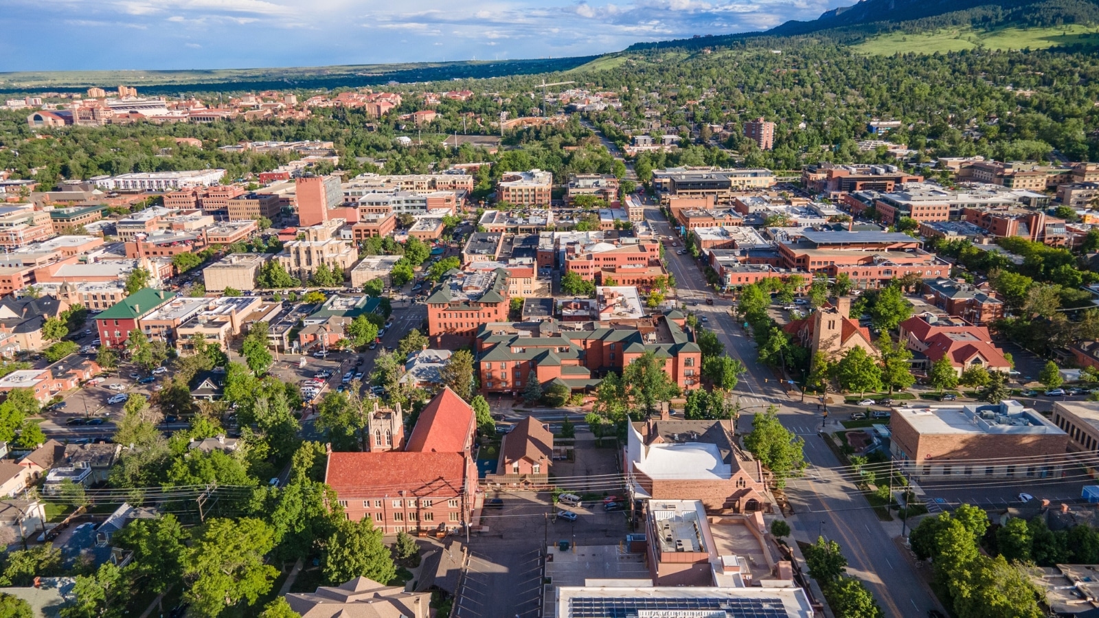 An aerial shot of downtown Boulder, Colorado, reveals a city with abundant greenery both within and around its limits. Streets are lined with greenery, contributing to a lush urban environment. The surrounding landscape includes rolling hills and open spaces, enhancing the natural beauty of the area.