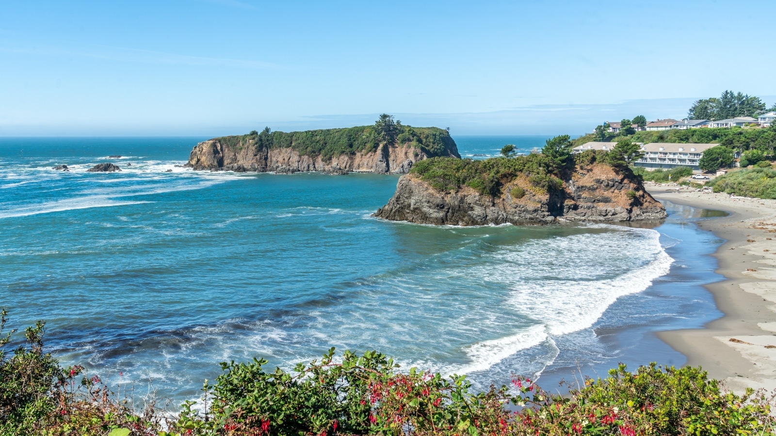 Brookings Beach features small waves and a medium-sized sandy shoreline. The water showcases oblong rock formations adorned with greenery.