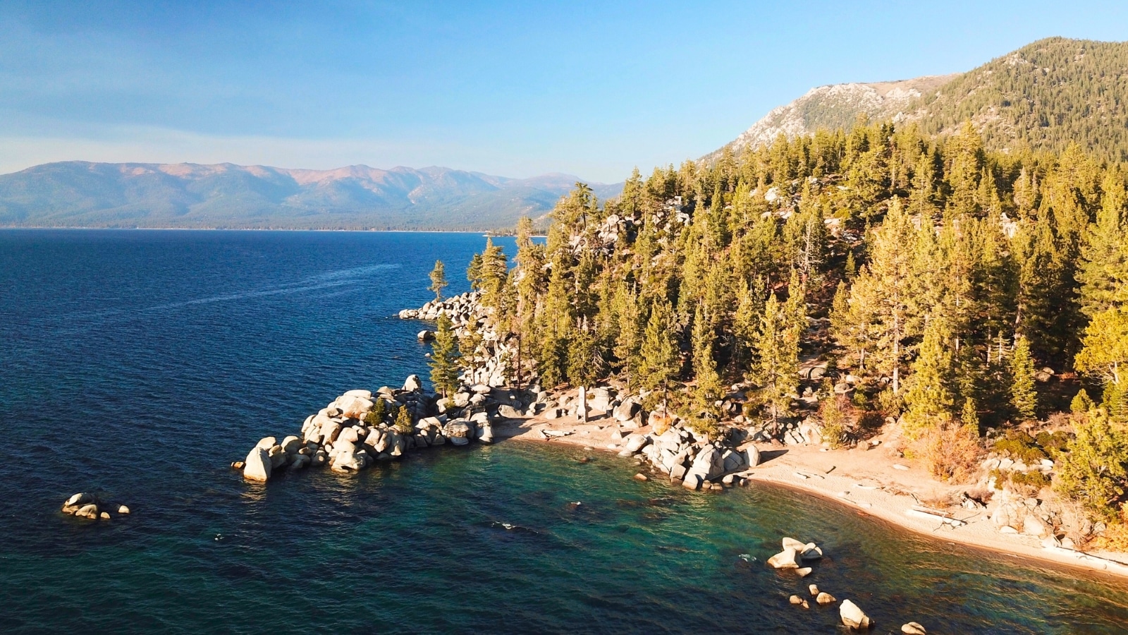 Chimney Beach on East Lake Tahoe features a narrow shoreline flanked by rocky terrain and a backdrop of dense forest. The hilly landscape adds a rugged charm to the beach setting. The lake itself presents a tranquil surface.