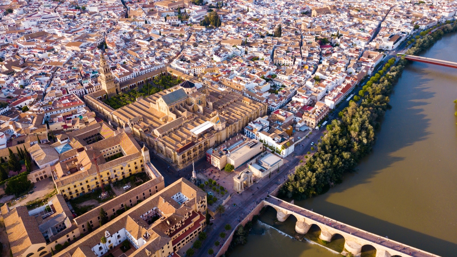 Historic Centre of Cordoba in Spain, featuring the antique Roman Bridge spanning the Guadalquivir River and the medieval Mosque-Cathedral. The Roman Bridge stands out with its ancient architectural style. Surrounding the area are buildings representing a blend of medieval and modern architecture.