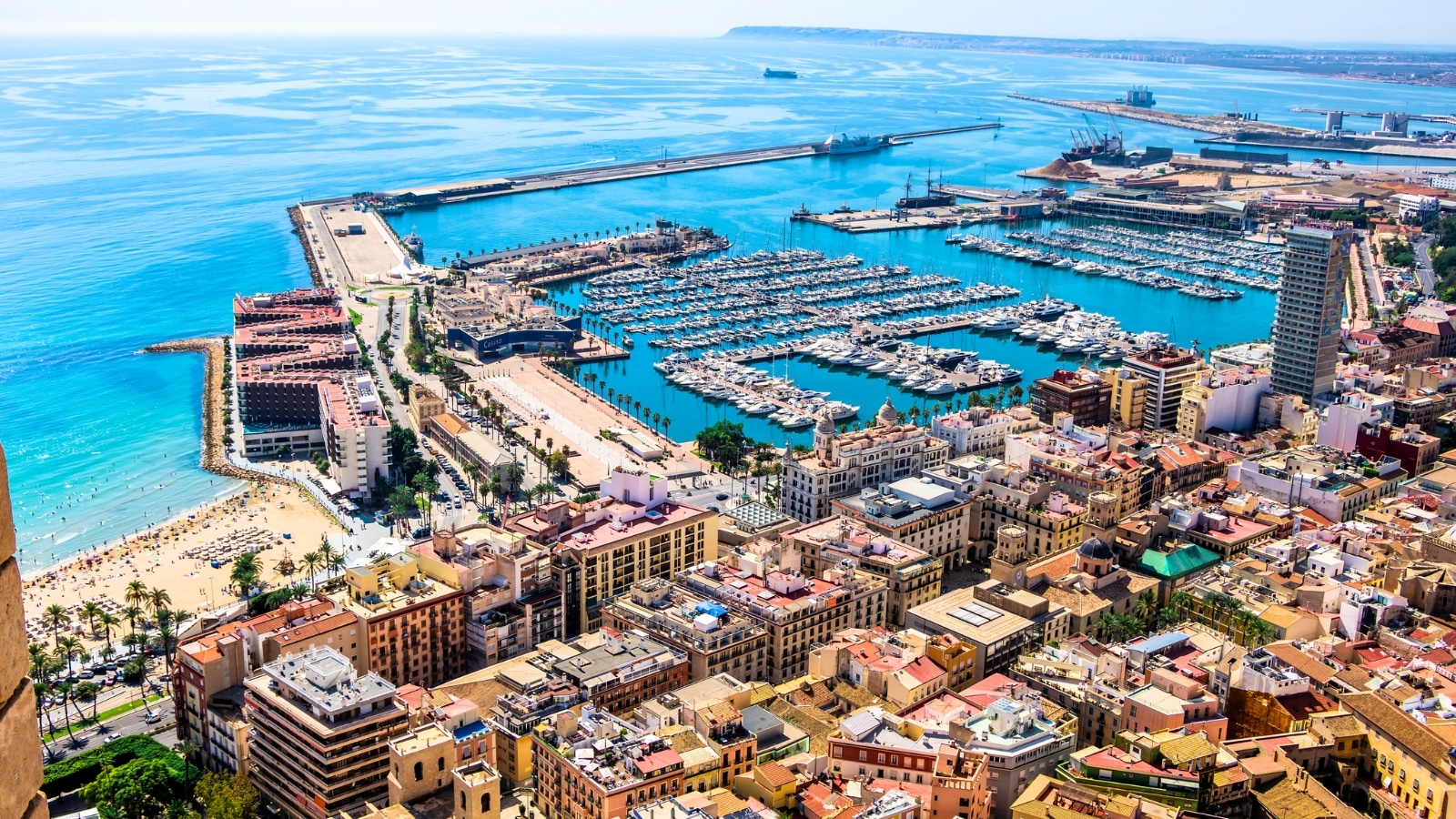 The beautiful port of Alicante, Spain, overlooks the Mediterranean Sea. This panoramic aerial view showcases the famous tourist city in Costa Blanca, Spain. The port consists of docks filled with boats and yachts, surrounded by waterfront promenades and colorful buildings.