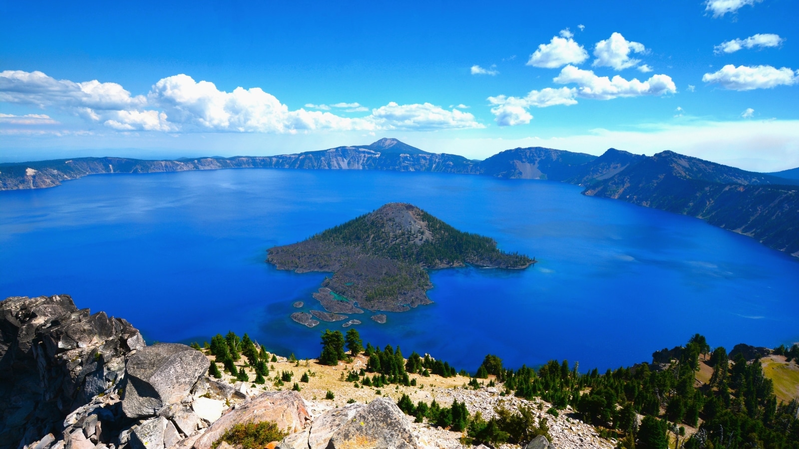 At the center of the lake is a hill adorned with greenery, providing a natural focal point. Surrounding the lake, rugged rocks frame the water.