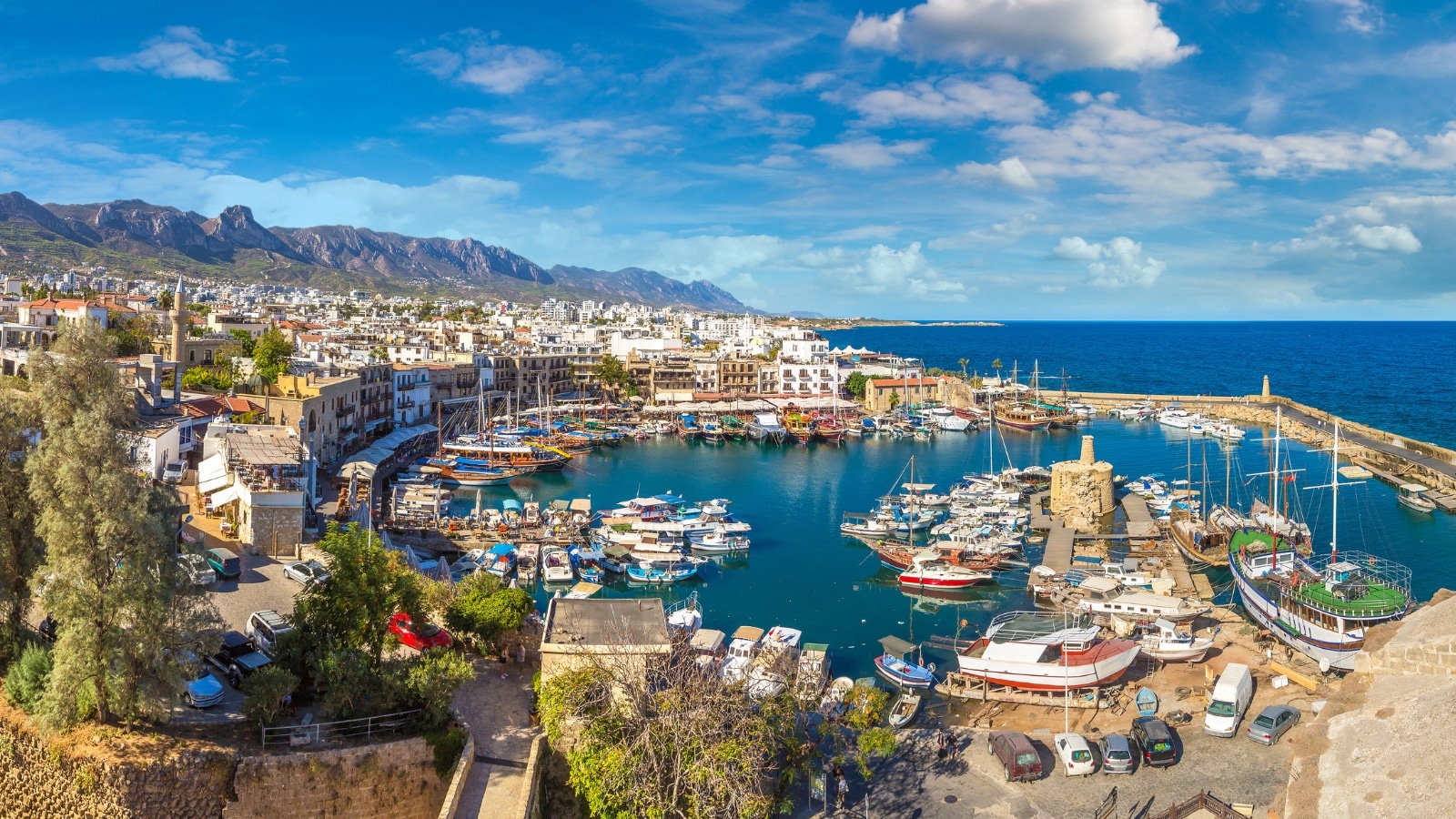 The image features a panoramic aerial view of the historic harbor in Kyrenia (Girne), located in North Cyprus, on a beautiful summer day. The harbor is characterized by a collection of boats and yachts of various sizes. Surrounding the harbor are buildings with traditional Mediterranean architecture.