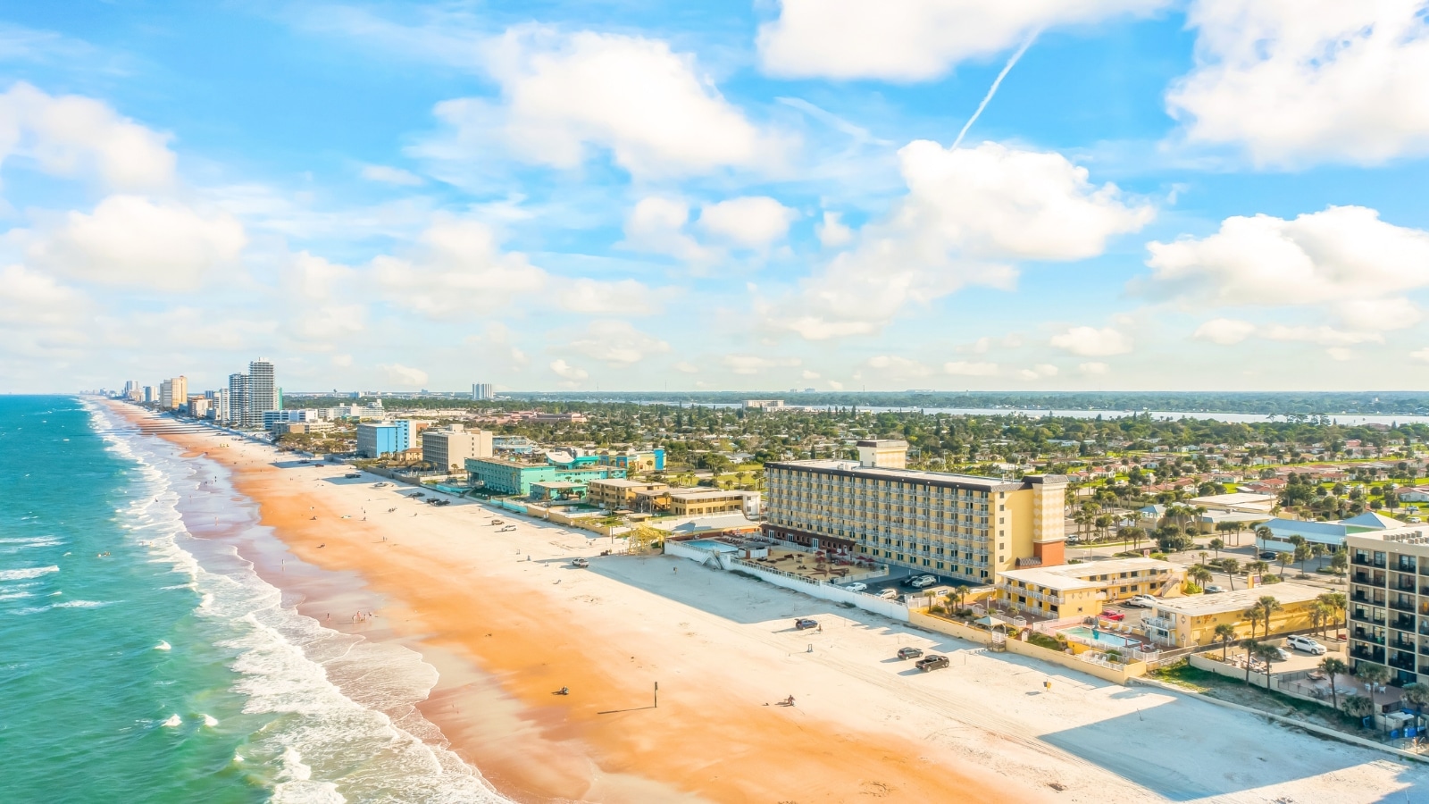 Drone photos of Daytona Beach, Florida, reveal beautiful beaches with striking blue water. The ocean lies to the left, with its deep, vibrant hues contrasting against the sandy shore. On the right, the beach extends into the city, where buildings and streets are visible. The beach itself is wide and sandy, with a mix of beachgoers and umbrellas dotting the shoreline.