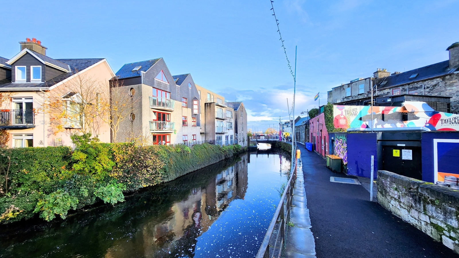 Galway, Ireland, with a river running through its center. On either side of the river are houses, predominantly beige in color, featuring gray triangular roofs.