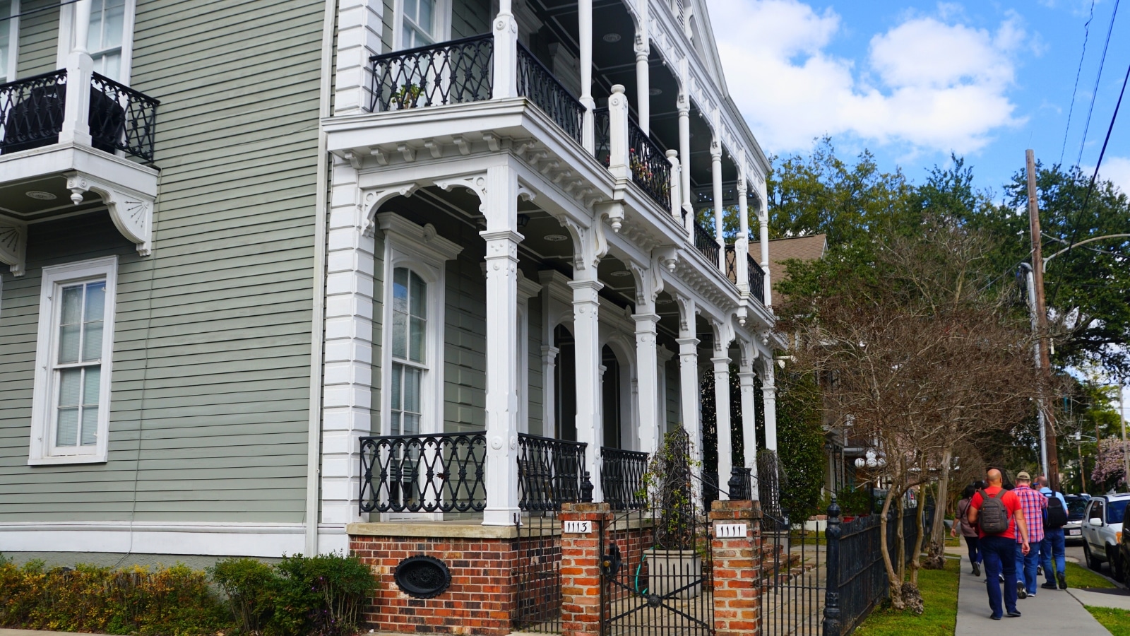 In New Orleans, Louisiana, tourists explore the beautiful houses of the Garden District on a walking tour. The scene includes a picturesque house and tourists walking along the sidewalk.