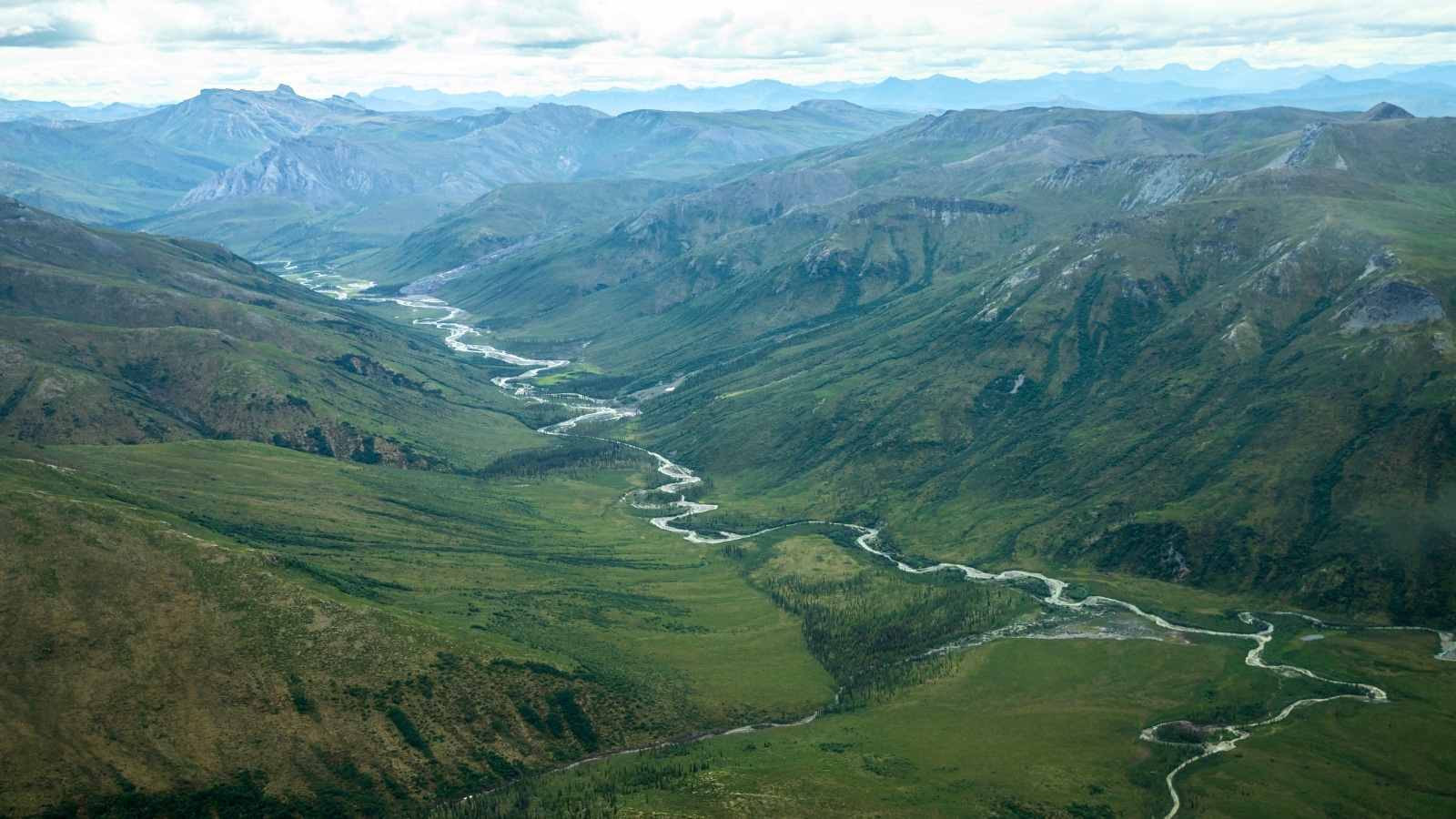 Gates of the Arctic National Park in Alaska features a landscape with hills and mountains flanking both sides. In the center, a winding river flows through the valley, surrounded by abundant greenery.