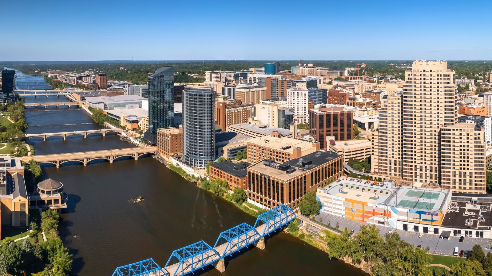 In Grand Rapids, Michigan, the city view includes a wide river crossed by six bridges in the foreground. The cityscape features tall buildings. Beyond the city, there is a significant amount of greenery visible, suggesting natural surroundings outside the urban center.