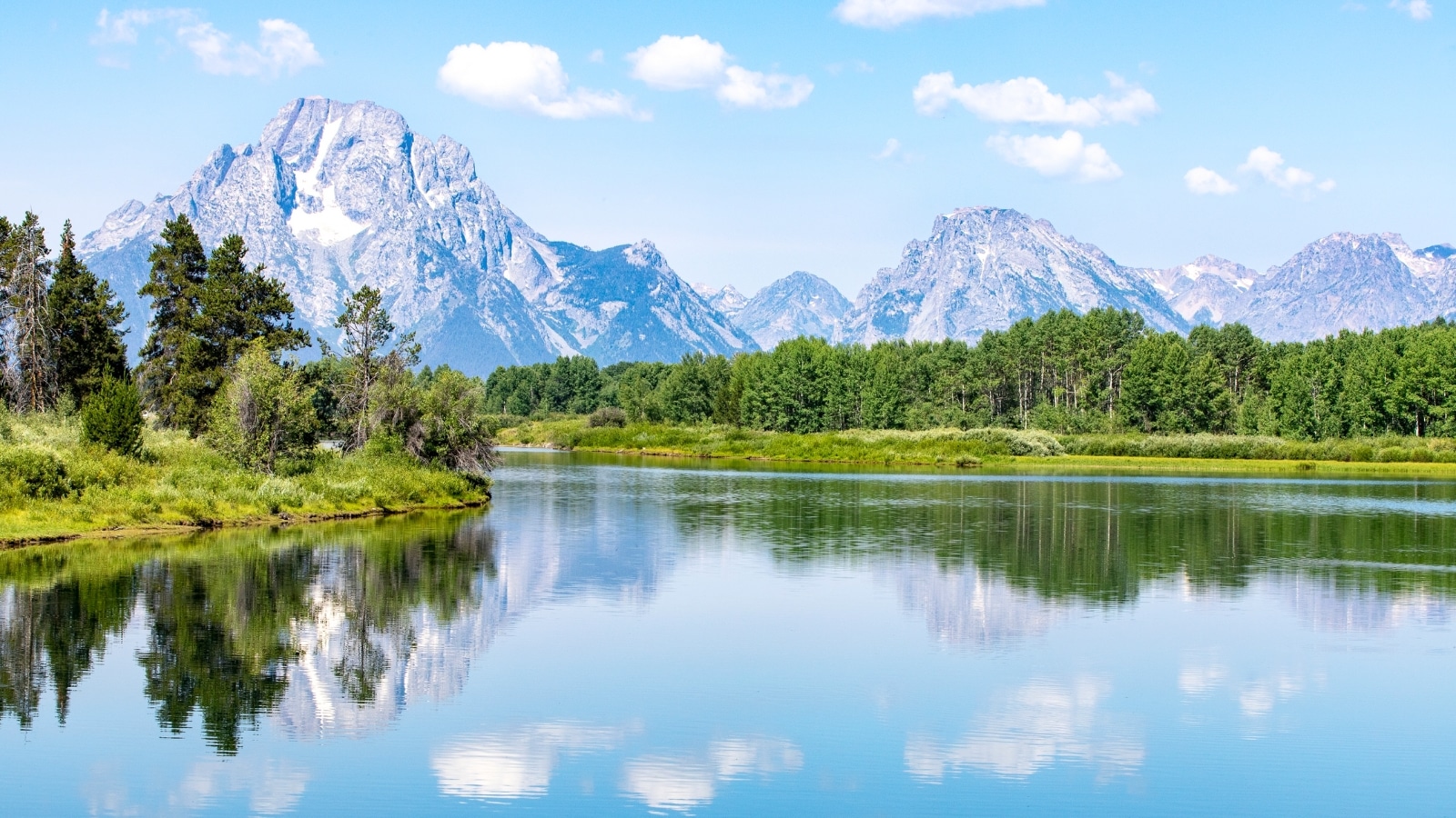 In this scene of Grand Teton National Park, Wyoming, a river flows in the foreground, surrounded by lush greenery typical of the park's landscape. The background features majestic rocky mountains, their peaks dusted with snow, creating a picturesque contrast against the vibrant greenery.