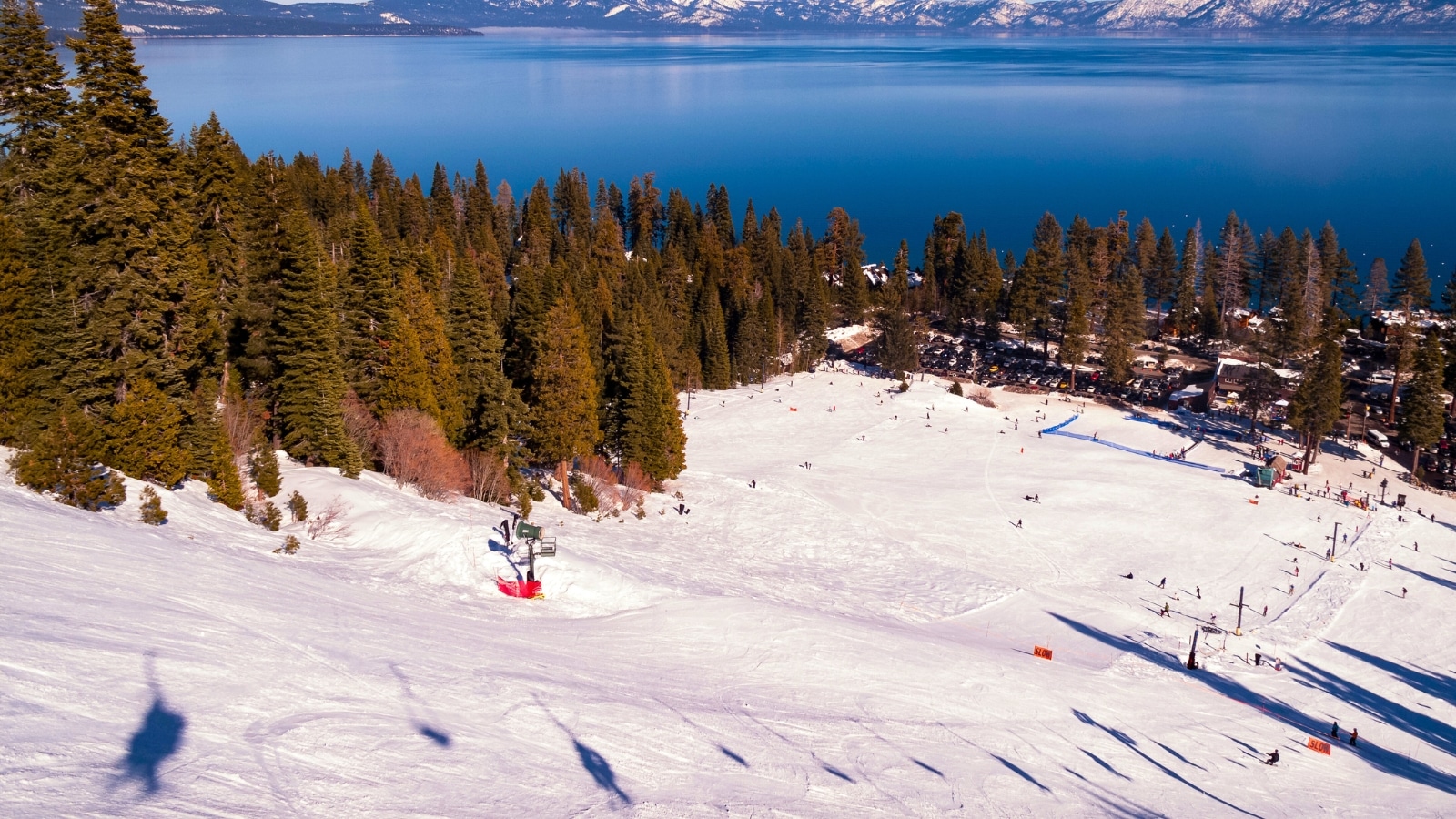 View of Lake Tahoe from a ski slope at Homewood Mountain Resort in California, USA. The scene features the vast expanse of the lake, surrounded by snow-covered slopes and coniferous trees typical of alpine environments. The color palette includes shades of white from the snow, various greens from the trees, and the deep blue of Lake Tahoe itself.