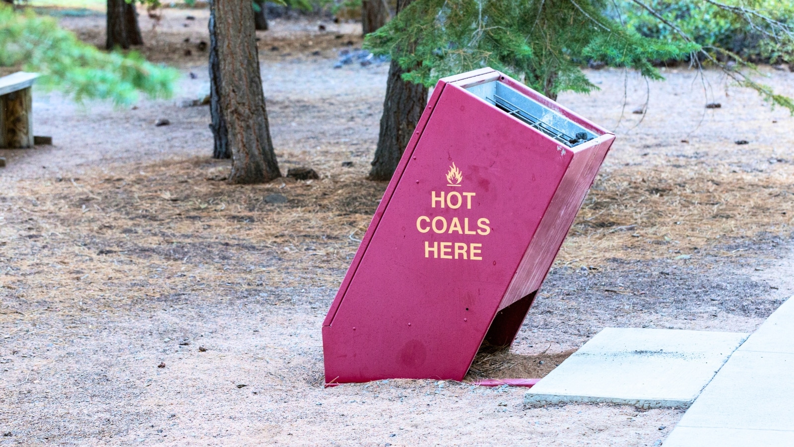 The image shows a hot coal disposal bin in a shaded day use area at Lake Tahoe. The bin appears sturdy and practical, designed to safely contain hot coals from grills or campfires.