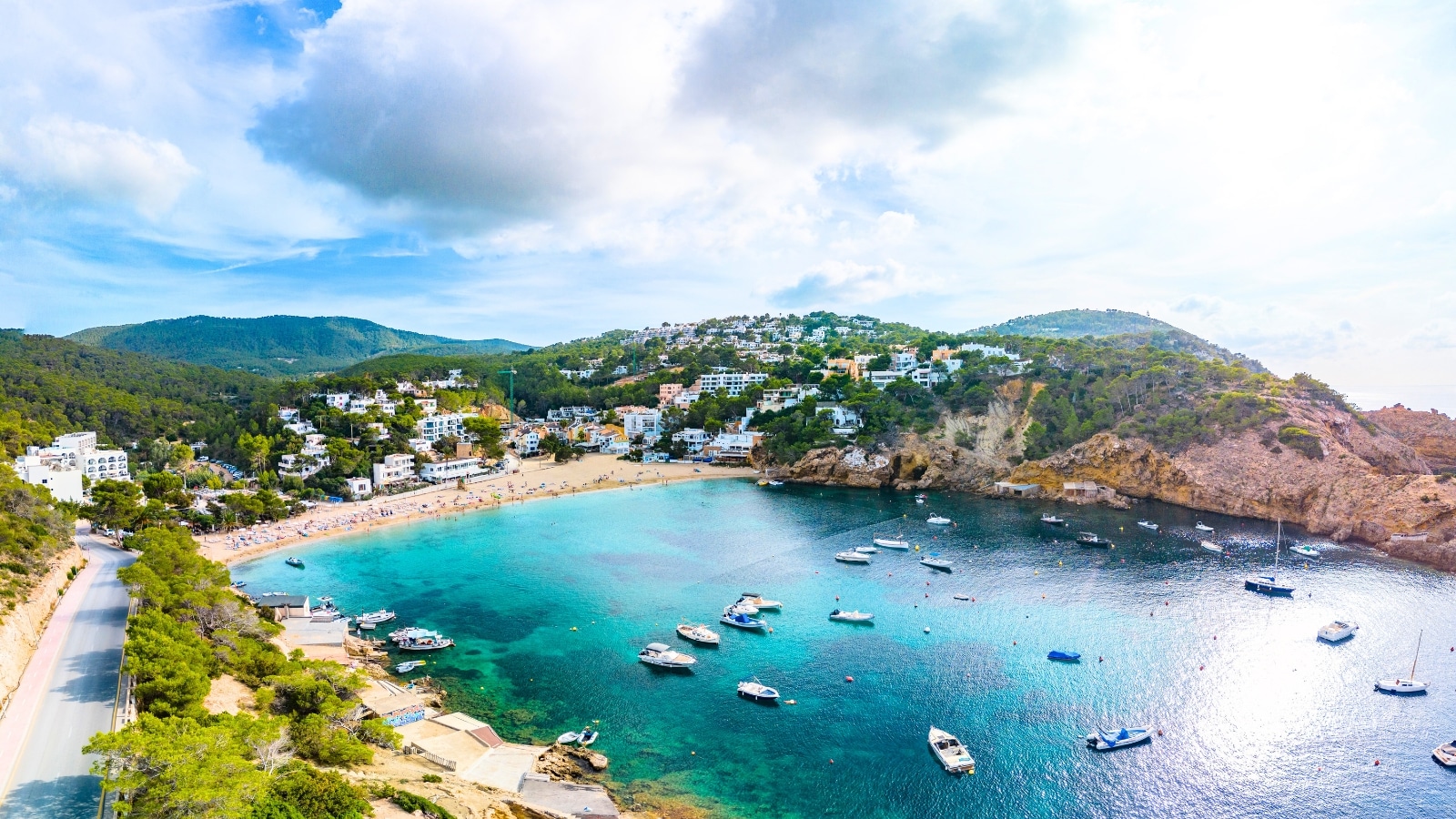 An aerial drone view captures Cala Vadella on the Ibiza islands in Spain. This coastal area features a crescent-shaped bay with sandy beaches and turquoise waters. The surrounding landscape includes rugged cliffs and lush greenery. The buildings near the bay are predominantly white. Boats and yachts dot the azure waters.